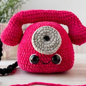 Può includere: Un telefono rosa all'uncinetto fatto a mano con un quadrante grigio e occhi neri. La cornetta è attaccata al corpo. Sono visibili anche un cavo nero e del filo rosa. Questo oggetto stravagante è un pezzo decorativo.