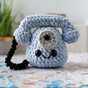 Puede incluir: Un teléfono de ganchillo azul claro con una cara sonriente, ojos negros y un dial gris. El auricular está conectado por un cable negro. El juguete está hecho de hilo suave y texturizado.