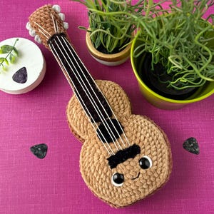 May include: A crocheted ukulele in tan and brown, featuring black strings and a smiling face. Two black guitar picks and a small white decorative item with a plant are also present. The ukulele is on a pink surface.