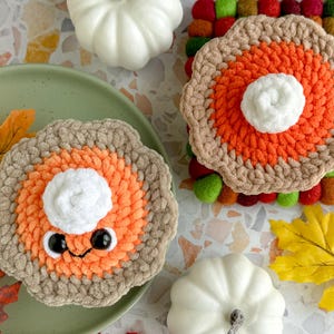 May include: Two crocheted pumpkin pie plush toys with smiling faces and googly eyes. The pies are decorated with orange and brown yarn and have white yarn for the whipped cream topping. The pies are sitting on a surface with fall-themed decorations.