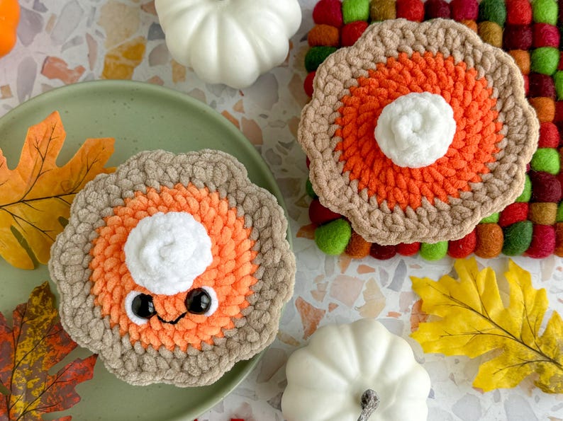 May include: Two crocheted pumpkin pie plush toys with smiling faces and googly eyes. The pies are decorated with orange and brown yarn and have white yarn for the whipped cream topping. The pies are sitting on a surface with fall-themed decorations.