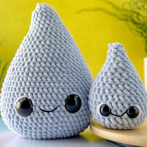Può includere: Due gocce di pioggia di peluche all'uncinetto di colore azzurro chiaro con occhi neri e un sorriso cucito. La goccia di pioggia più grande è in primo piano e quella più piccola è dietro.