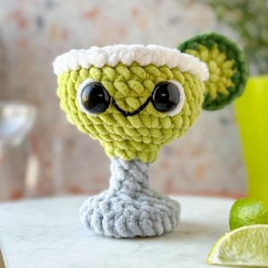 May include: A green crocheted margarita glass with a white rim and googly eyes. The glass has a lime wedge on the side and is sitting on a white surface.