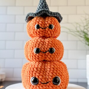 Pu&ograve; includere: Una pila di tre zucche arancioni all'uncinetto con occhi neri e bocche sorridenti. La zucca in cima indossa un cappello da strega grigio. Le zucche sono impilate, creando una stravagante decorazione di Halloween.