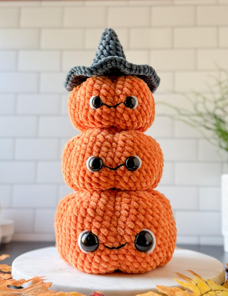 Pu&ograve; includere: Una pila di tre zucche arancioni all'uncinetto con occhi neri e bocche sorridenti. La zucca in cima indossa un cappello da strega grigio. Le zucche sono impilate, creando una stravagante decorazione di Halloween.