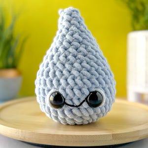 Può includere: Un giocattolo di peluche a forma di goccia di pioggia di colore azzurro chiaro lavorato a uncinetto con occhi neri e un sorriso cucito. Il giocattolo è seduto su una superficie di legno.