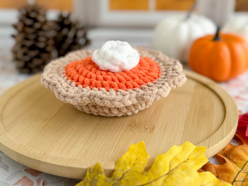 May include: A crocheted pumpkin pie with a brown crust, orange filling, and white whipped cream topping. The pie is sitting on a wooden plate with fall leaves around it.