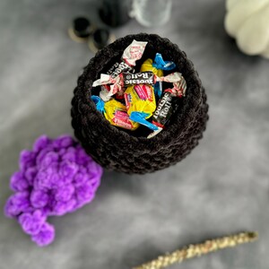 K&ouml;nnte beinhalten: Ein schwarzer geh&auml;kelter Kessel gef&uuml;llt mit verschiedenen S&uuml;&szlig;igkeiten, darunter Tootsie Rolls und andere verpackte S&uuml;&szlig;igkeiten. Der Kessel steht auf einer grauen Oberfl&auml;che.