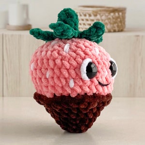 Peut inclure: Un jouet en peluche en forme de fraise crochet&eacute; rose et marron avec une feuille crochet&eacute; verte et des pois blancs. Le jouet a un visage souriant avec des yeux noirs et une bouche blanche.