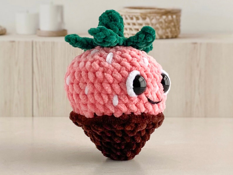 Peut inclure: Un jouet en peluche en forme de fraise crochet&eacute; rose et marron avec une feuille crochet&eacute; verte et des pois blancs. Le jouet a un visage souriant avec des yeux noirs et une bouche blanche.