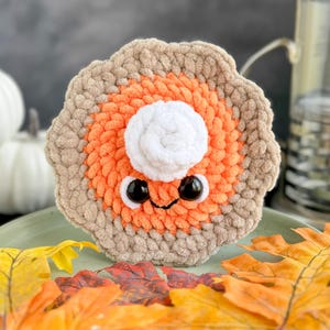 May include: A crocheted pumpkin pie plush toy with a white whipped cream topping and googly eyes. The pie is sitting on a green plate with fall leaves.