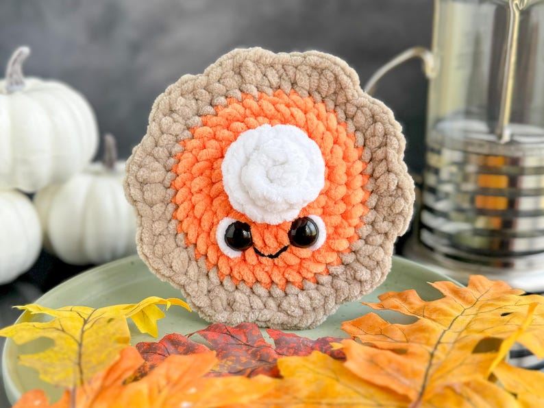 May include: A crocheted pumpkin pie plush toy with a white whipped cream topping and googly eyes. The pie is sitting on a green plate with fall leaves.