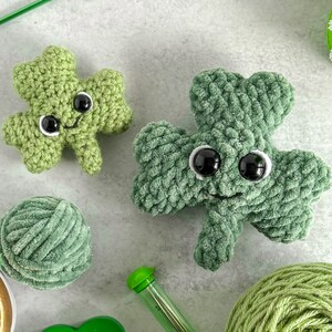 May include: Two green crocheted shamrock plushies with black eyes and a smiling mouth. The plushies are sitting on a gray surface with yarn and a crochet hook.