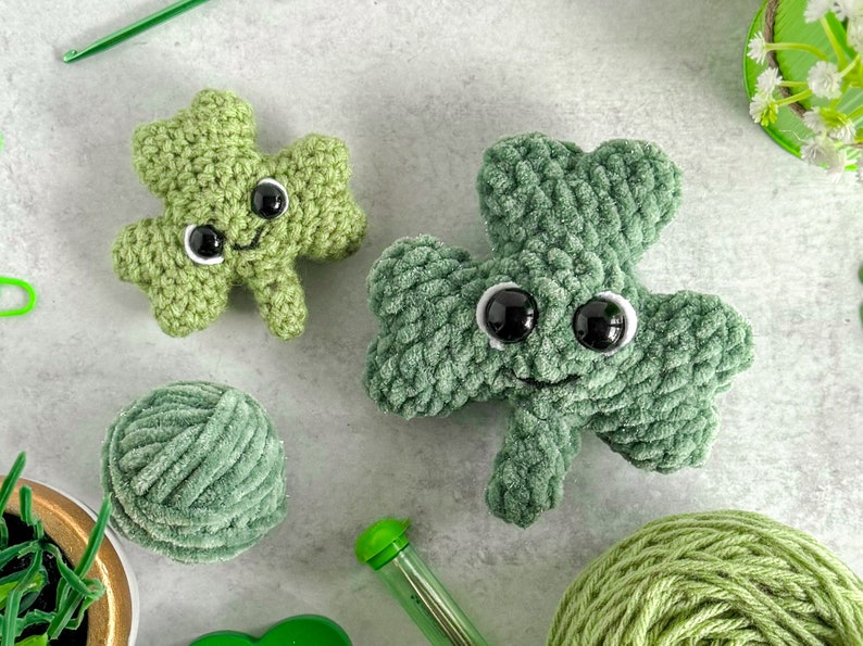 May include: Two green crocheted shamrock plushies with black eyes and a smiling mouth. The plushies are sitting on a gray surface with yarn and a crochet hook.