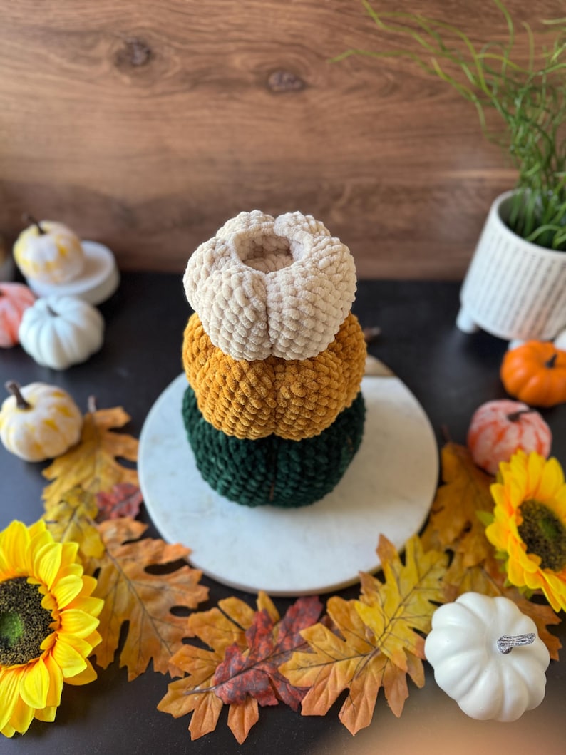Pu&ograve; includere: Una pila di tre zucche di peluche fatte a mano in crema, oro e verde. Le zucche sono disposte su un piatto di marmo bianco, circondate da foglie autunnali, girasoli e piccole zucche decorative. Una decorazione autunnale.