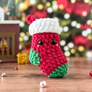 Puede incluir: Un calcetín de Navidad de ganchillo hecho a mano en rojo, blanco y verde. El calcetín tiene un puño blanco y detalles verdes en la punta y el talón. Cuenta con ojos de seguridad negros y un pequeño lazo para colgar. Decoración festiva navideña.