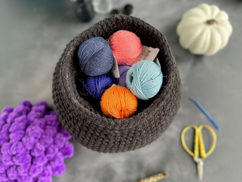 Pu&ograve; includere: Un cesto di uncinetto grigio pieno di gomitoli di lana colorati. I gomitoli di lana sono in tonalit&agrave; di blu, arancione, rosa e viola.
