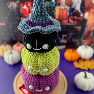 Pu&ograve; includere: Una pila di tre zucche all'uncinetto nere, verdi e viola, sormontata da un cappello da strega blu con finiture viola. Ogni zucca ha una faccia sorridente con occhi bianchi. La pila &egrave; su un piatto di legno.