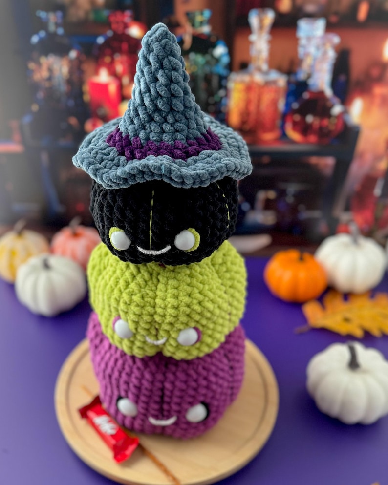 Pu&ograve; includere: Una pila di tre zucche all'uncinetto nere, verdi e viola, sormontata da un cappello da strega blu con finiture viola. Ogni zucca ha una faccia sorridente con occhi bianchi. La pila &egrave; su un piatto di legno.