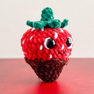Peut inclure: Un jouet en peluche en forme de fraise crochet&eacute; avec un fond enrob&eacute; de chocolat. La fraise a une feuille crochet&eacute; verte, des yeux blancs et une bouche souriante.