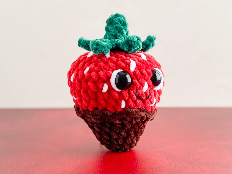 Peut inclure: Un jouet en peluche en forme de fraise crochet&eacute; avec un fond enrob&eacute; de chocolat. La fraise a une feuille crochet&eacute; verte, des yeux blancs et une bouche souriante.