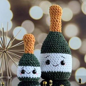 May include: Two crocheted champagne bottle plush toys with smiling faces. The larger bottle is green and white, and the smaller bottle is green and orange.