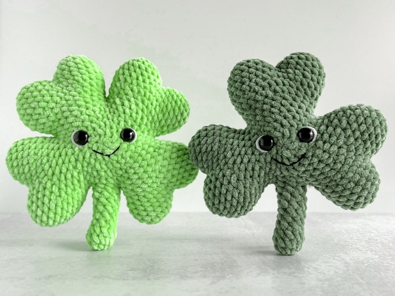 May include: Two crocheted plush shamrock toys with smiling faces and black eyes. One shamrock is green and the other is a darker green.