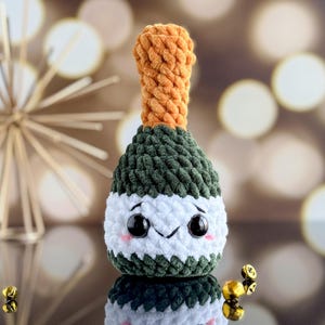 May include: A green and white crocheted champagne bottle with a cute cartoon face. The bottle has a brown crocheted top and is sitting on a reflective surface.
