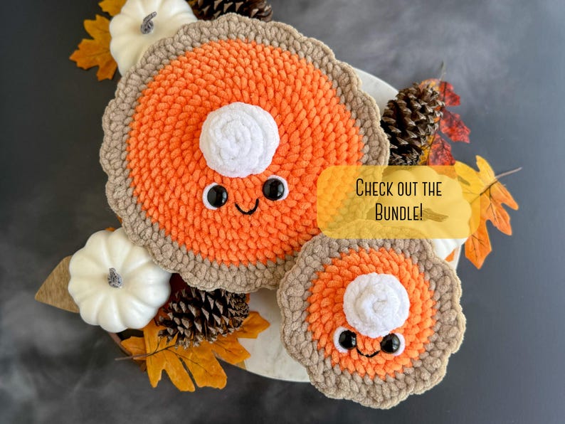 May include: Two crocheted pumpkin pie plush toys with smiling faces and white whipped cream topping. The larger pie is orange and brown, the smaller pie is orange and brown. The pies are on a white surface with fall leaves and pine cones.