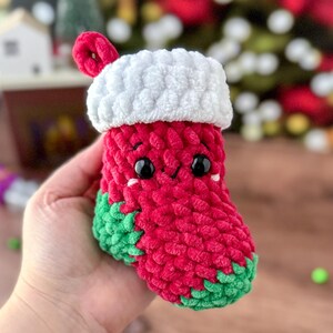 K&ouml;nnte beinhalten: Eine handgefertigte, rot-gr&uuml;ne H&auml;kel-Weihnachtsstrumpf mit wei&szlig;er Manschette und Schlaufe. Der Strumpf hat schwarze Augen und ein l&auml;chelndes Gesicht. Der Strumpf wird in einer Hand gehalten.