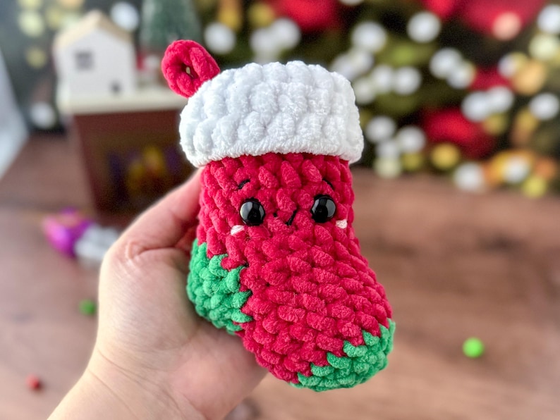 K&ouml;nnte beinhalten: Eine handgefertigte, rot-gr&uuml;ne H&auml;kel-Weihnachtsstrumpf mit wei&szlig;er Manschette und Schlaufe. Der Strumpf hat schwarze Augen und ein l&auml;chelndes Gesicht. Der Strumpf wird in einer Hand gehalten.