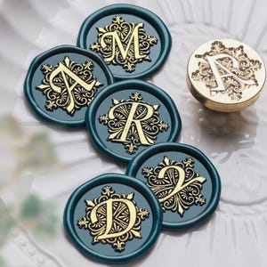 May include: A collection of teal wax seals with gold embossed letters and decorative flourishes. The seals are arranged on a white surface, with a gold metal stamp featuring the letter "A" visible.