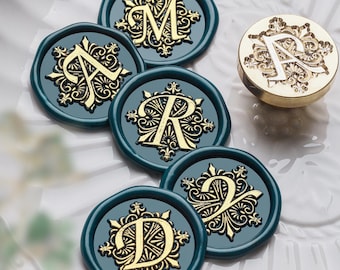 May include: A collection of teal wax seals with gold embossed letters and decorative flourishes. The seals are arranged on a white surface, with a gold metal stamp featuring the letter "A" visible.