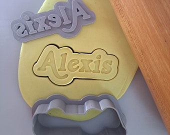 May include: A gray plastic cookie cutter in the shape of the name "Alexis" with a rolling pin and yellow fondant in the background.