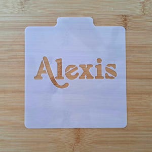 May include: White plastic stencil with the name "Alexis" cut out in a cursive font.