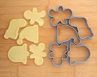 Puede incluir: Un juego de cortadores de galletas de Navidad y las formas cortadas de galletas sobre una superficie de madera. Los cortadores son grises y las galletas son de color amarillo claro. Los cortadores incluyen una campana, una media, una estrella, un reno y un muñeco de nieve. Las galletas incluyen una campana, una media, una estrella, un reno y un muñeco de nieve.