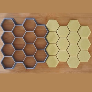 May include: A gray plastic honeycomb cookie cutter next to a sheet of yellow cookie dough cut into hexagonal shapes.
