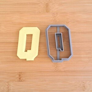 May include: A gray plastic cookie cutter in the shape of the number 0, with a yellow plastic cookie in the same shape next to it.