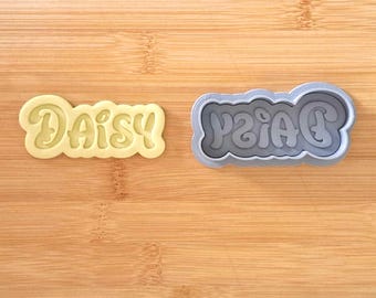 May include: A gray plastic cookie cutter in the shape of the word "Daisy" with a yellow cookie cut out using the cutter.