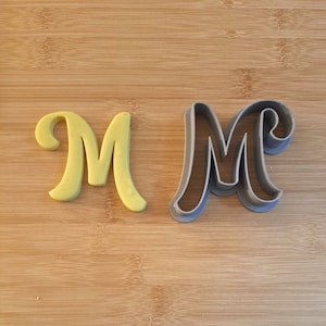 Puede incluir: Un cortador de galletas de plástico amarillo en forma de letra M y un cortador de galletas de plástico gris en forma de letra M sobre una superficie de madera.