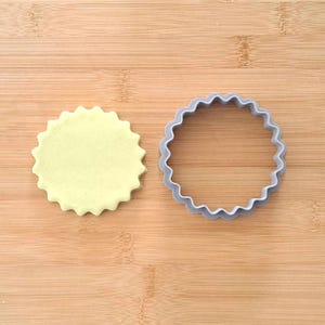 May include: A light green cookie dough disc next to a gray plastic cookie cutter with a scalloped edge.