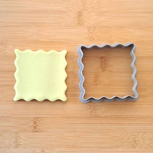 May include: A light green square of fondant dough next to a gray plastic cookie cutter with a wavy edge.