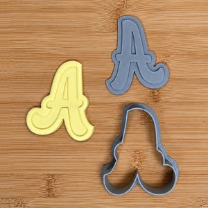 May include: Three letter A cookie cutters. One is a solid yellow plastic shape, while the other two are grey plastic outlines. The grey cutters show both the top profile and the bottom cutting edge, while the yellow cutter shows the top face.