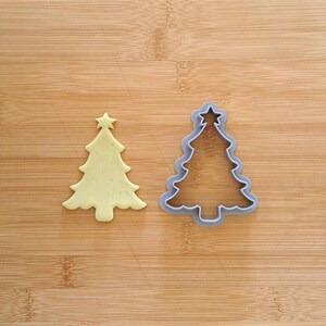 Op de afbeelding: Een grijze plastic kerstboomkoekjesvormpje naast een gele kerstboomkoekje op een houten oppervlak.