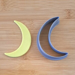 Puede incluir: Dos cortadores de galletas en forma de media luna, uno amarillo y otro morado, sobre una superficie de madera.