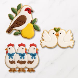 May include: Assortment of decorated sugar cookies in the shape of a bird with a pear, two doves, and three chickens. The cookies are white, brown, red, yellow, and green, with intricate details and a smooth finish. The cookies are arranged on a white surface.