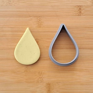 May include: A gray plastic cookie cutter shaped like a teardrop, next to a yellow teardrop-shaped cookie.