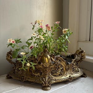 May include: An antique brass planter, filled with green foliage and delicate pink and cream flowers. The planter has ornate scrollwork, cherub figures, and a detailed, aged finish. A decorative piece for home decor.