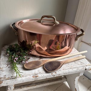 May include: A copper oval casserole pot with a lid and brass handles sits on a distressed white wooden stool. Two wooden spoons and a sprig of herbs with purple flowers are next to the pot. The pot has a shiny, reflective surface.