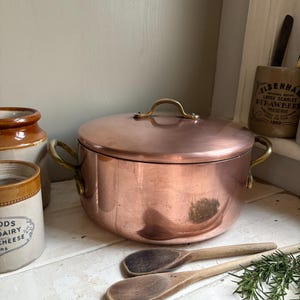 May include: A copper cooking pot with a lid and brass handles. Two wooden spoons are in front of the pot. The pot is round and has a shiny surface. Other ceramic jars are visible in the background.